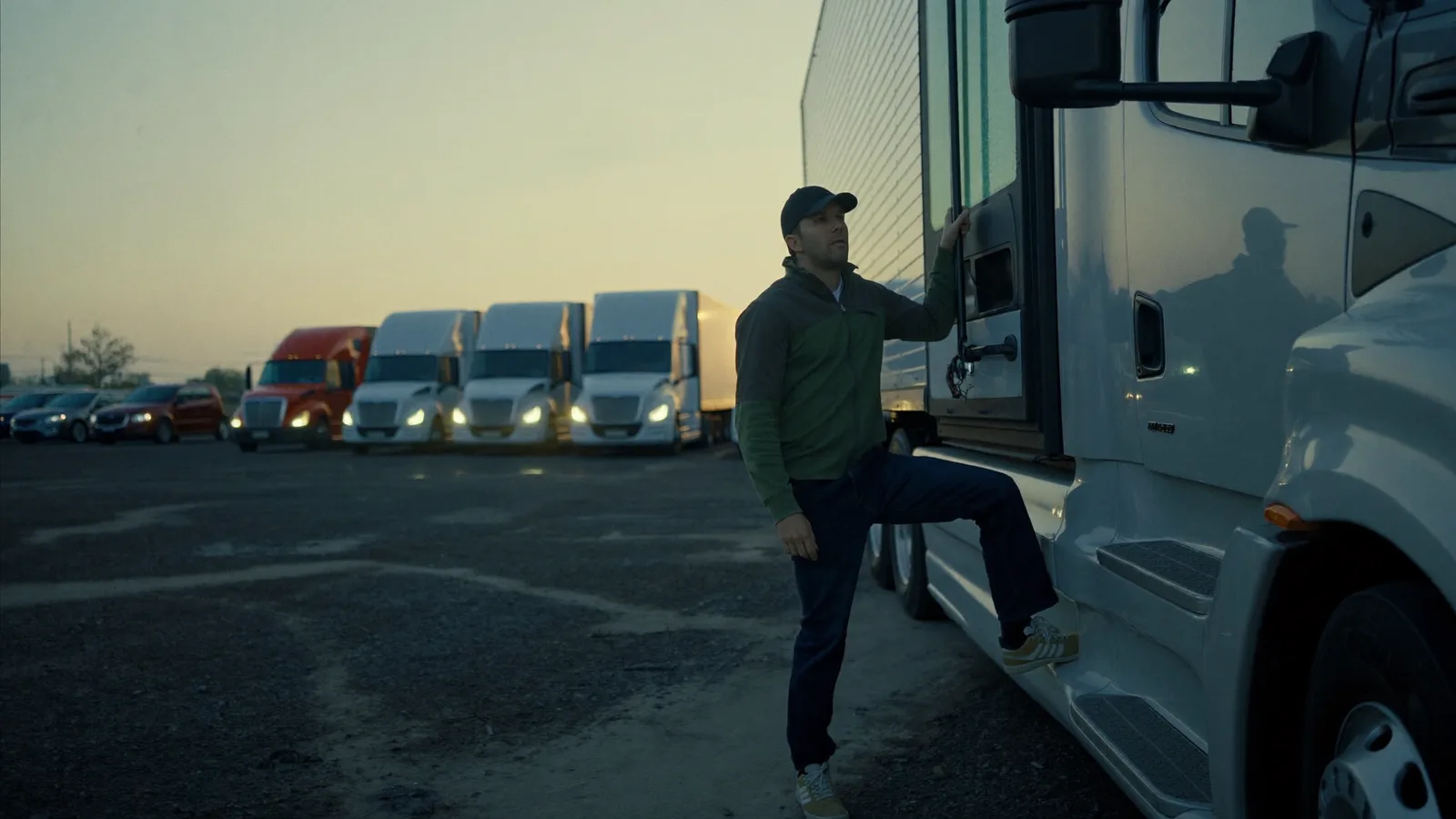 Dispatcher working through load planning at a desk in a small trucking office at first light, trucks visible in the yard outside.