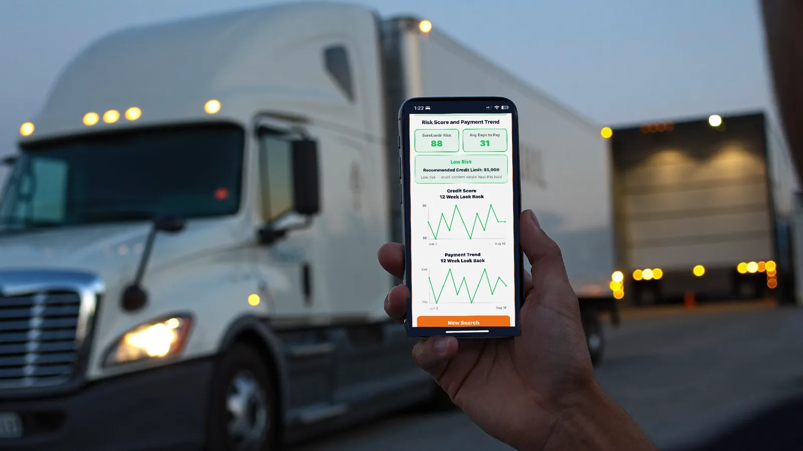 A carrier holding a phone showing a SureLoadr freight broker report with risk score and days to pay, a truck and loading dock blurred behind at dusk.
