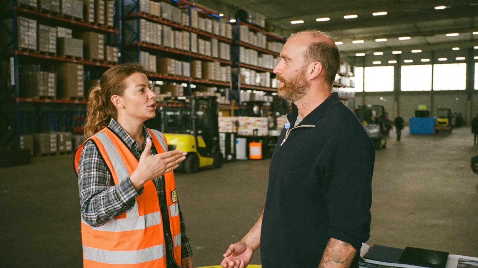 A shipping manager in a hi-vis vest talking with a freight broker on a busy warehouse floor, forklifts and loaded racking behind them.